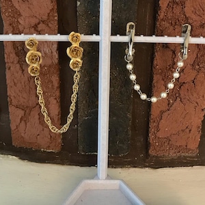 May include: Two pairs of earrings on a white display stand. The first pair features a gold chain with three gold rose charms. The second pair features a silver chain with white pearl charms.