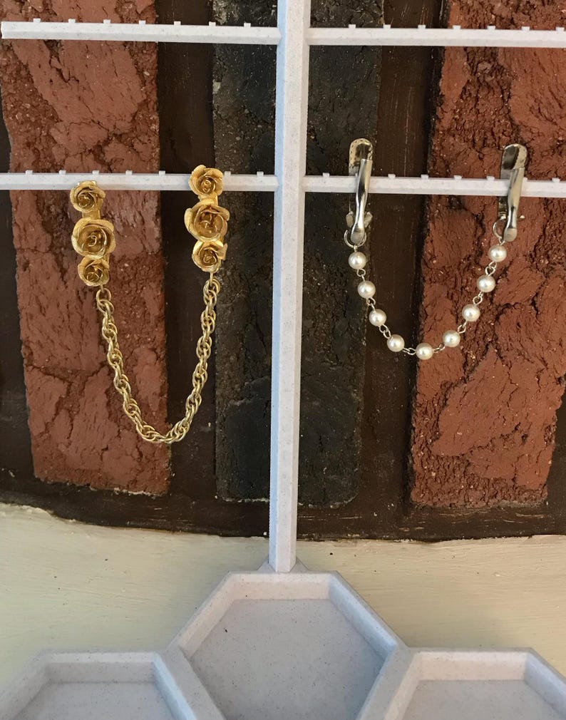 May include: Two pairs of earrings on a white display stand. The first pair features a gold chain with three gold rose charms. The second pair features a silver chain with white pearl charms.