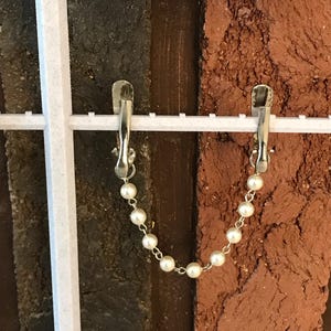 May include: Two silver clips with a white pearl chain hanging from each. The clips are attached to a white grid background with a brown brick wall behind.
