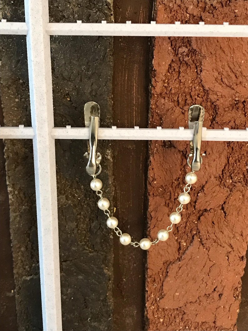 May include: Two silver clips with a white pearl chain hanging from each. The clips are attached to a white grid background with a brown brick wall behind.