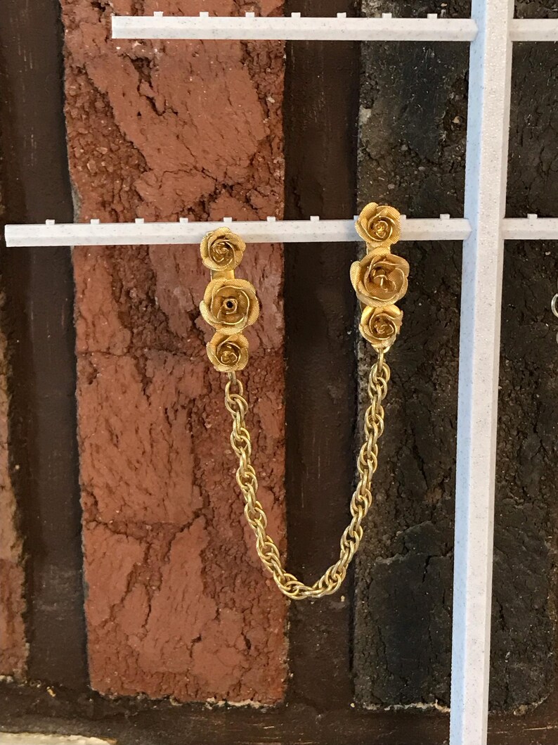 May include: A pair of gold-tone earrings with a chain connecting them. Each earring features three small rose-shaped charms.