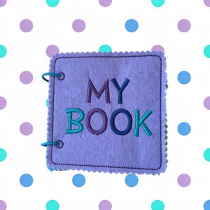 May include: A lavender felt book with the words "MY BOOK" embroidered in teal and purple. The book has a scalloped edge and metal rings for binding. The background is white with teal, purple, and blue polka dots.