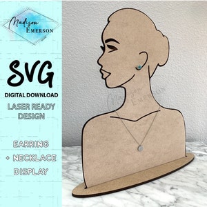 May include: A wooden earring and necklace display stand in the shape of a woman's head. The stand is brown and has a cutout of a woman's face. The stand has a small round base. The stand is designed to hold earrings and a necklace.
