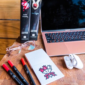 May include: A white notepad with a red and black illustration of a bouquet of flowers. Three red and black markers are on a wooden surface next to the notepad. Two books titled "New Moon" and "Twilight" are in the background.