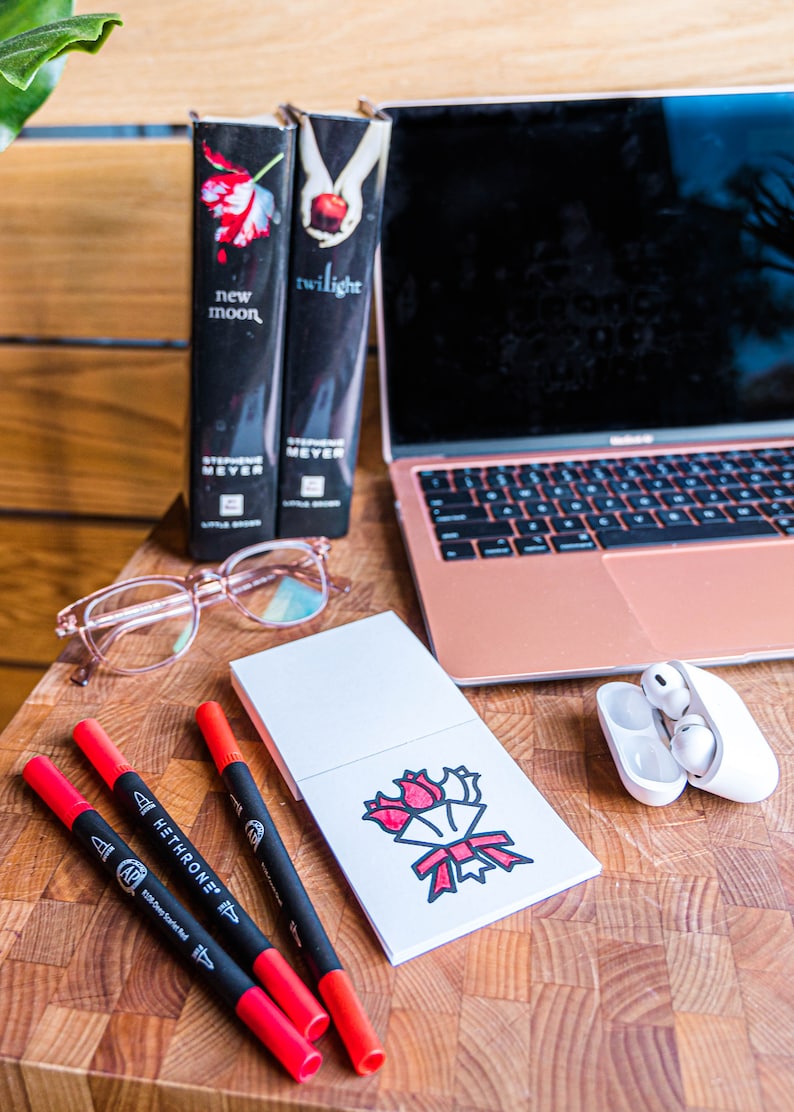 May include: A white notepad with a red and black illustration of a bouquet of flowers. Three red and black markers are on a wooden surface next to the notepad. Two books titled "New Moon" and "Twilight" are in the background.