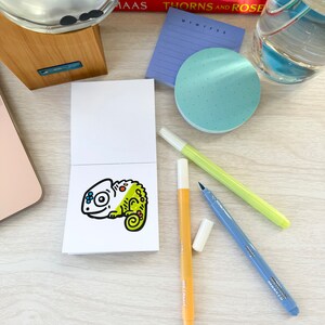 Op de afbeelding: Een klein wit notitieboekje met een cartoon-kameleonillustratie op de voorkant. De kameleon is groen, geel, blauw en rood. Er liggen drie stiften op tafel: een gele stift, een blauwe stift en een oranje stift.