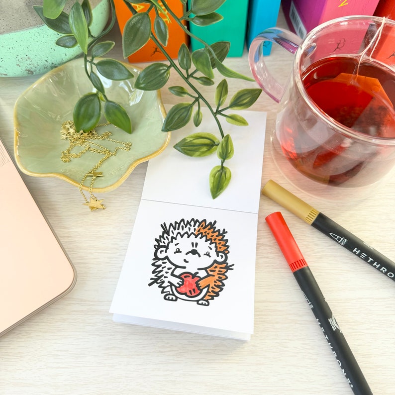 Op de afbeelding: Een handgetekend ontwerp van een egel die een rood hart vasthoudt. De egel is getekend in zwart en wit met oranje en bruine accenten. De illustratie is op een witte kaart.