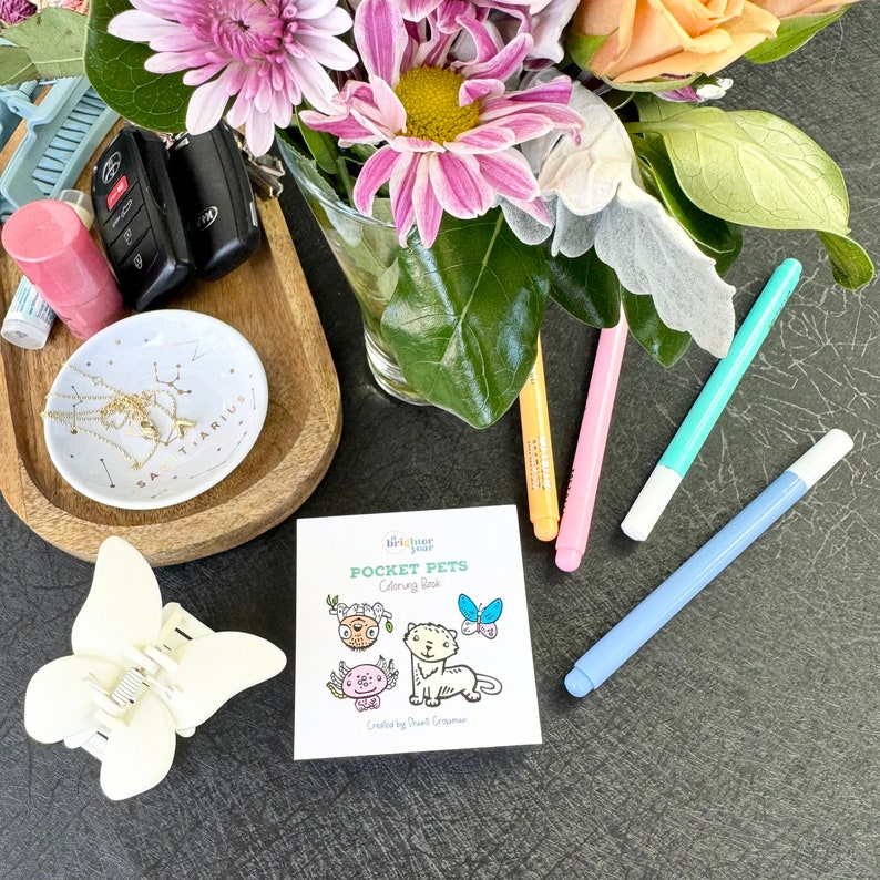 Op de afbeelding: Een klein kleurboek met de titel "Pocket Pets Coloring Book" van "Little Blue Year" met illustraties van een kat, een vlinder, een kikker en een schildpad. Het boek ligt op een zwart oppervlak met verschillende kleurpotloden en andere voorwerpen.