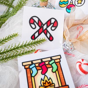 May include: Two square white cards featuring colorful holiday illustrations. One card displays two crossed red and white candy canes. The other card shows a fireplace with a burning fire and three hanging stockings in blue, red, and green.