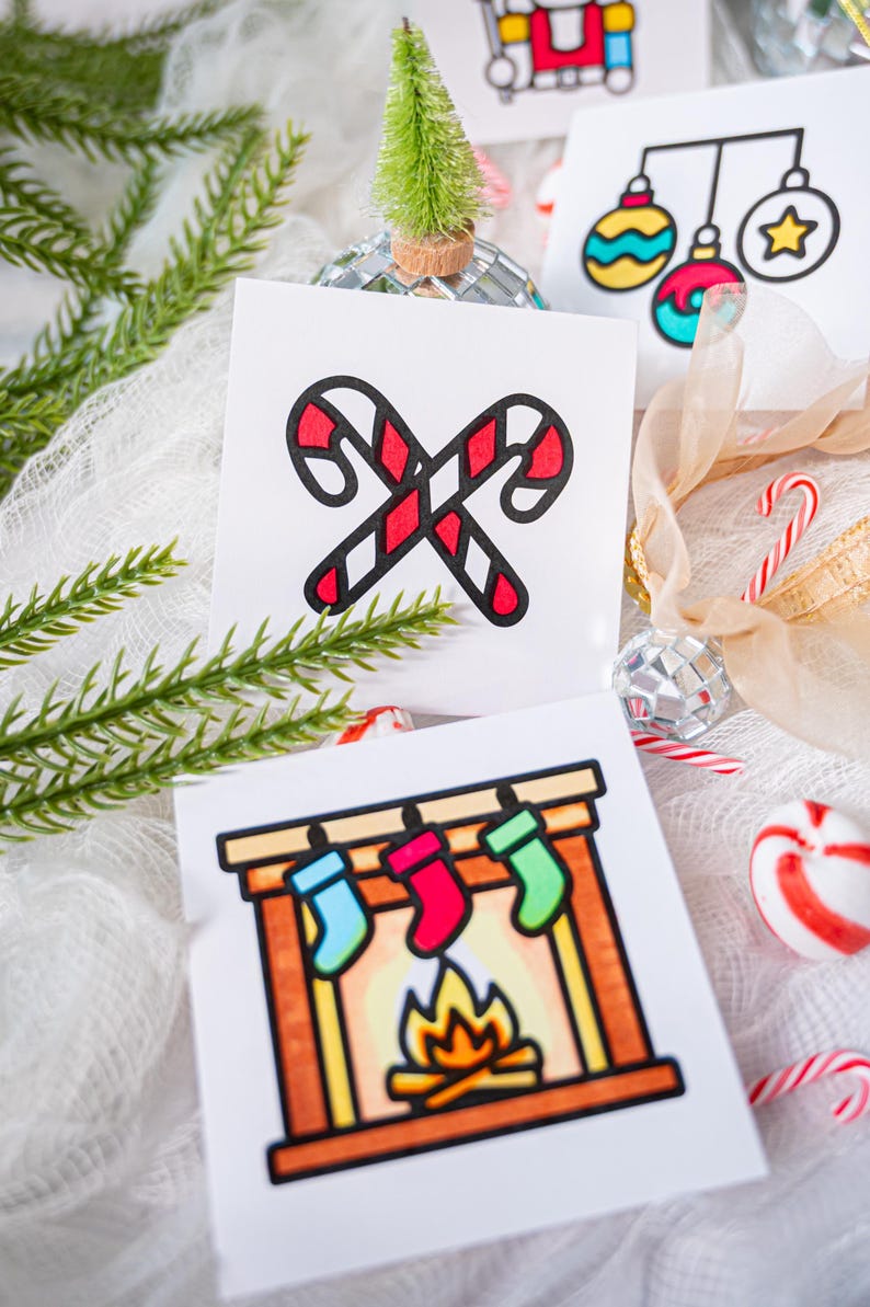 May include: Two square white cards featuring colorful holiday illustrations. One card displays two crossed red and white candy canes. The other card shows a fireplace with a burning fire and three hanging stockings in blue, red, and green.