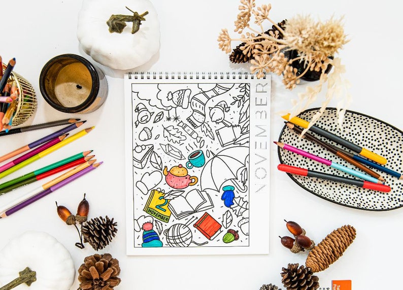 May include: A coloring book with a November theme featuring a variety of fall-themed illustrations, including leaves, acorns, a teapot, an umbrella, and a book. The book is open to a page with the date "2 November" written on it. The book is surrounded by colored pencils, pine cones, and other fall decorations.
