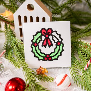 May include: A square white card featuring a centered graphic illustration of a green holly wreath with a red bow and red berries, outlined in thick black lines. The design is simple, flat, and uses bold colors to depict the holiday foliage.