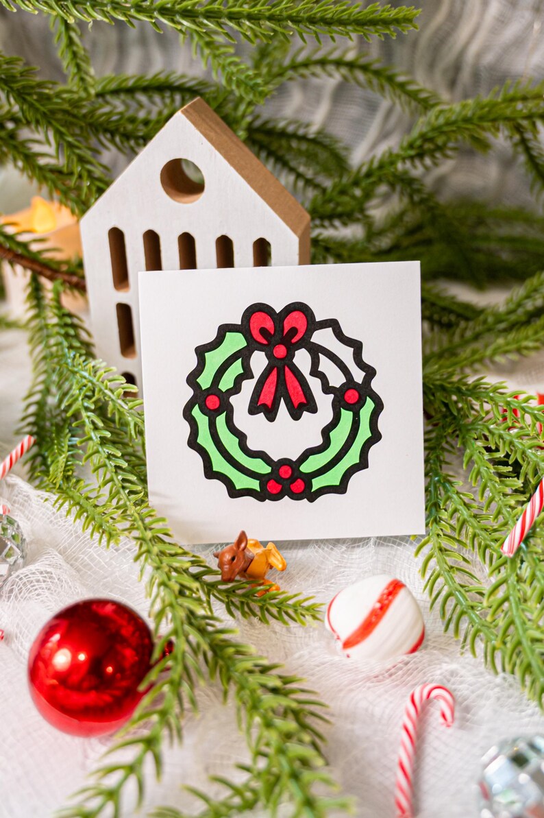 May include: A square white card featuring a centered graphic illustration of a green holly wreath with a red bow and red berries, outlined in thick black lines. The design is simple, flat, and uses bold colors to depict the holiday foliage.