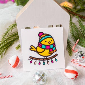 May include: A square white card featuring a cartoon illustration of a yellow bird wearing a teal and magenta striped winter hat and scarf. The bird is perched on a brown branch decorated with a string of small, colorful, glowing holiday lights.