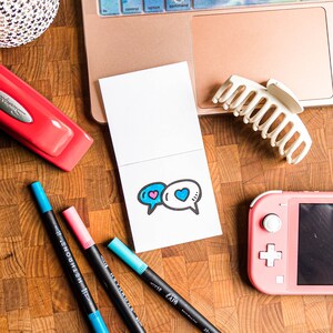 May include: A white card with a blue and pink heart design inside two speech bubbles. The card is on a wooden surface with a laptop, a stapler, and a few pens.