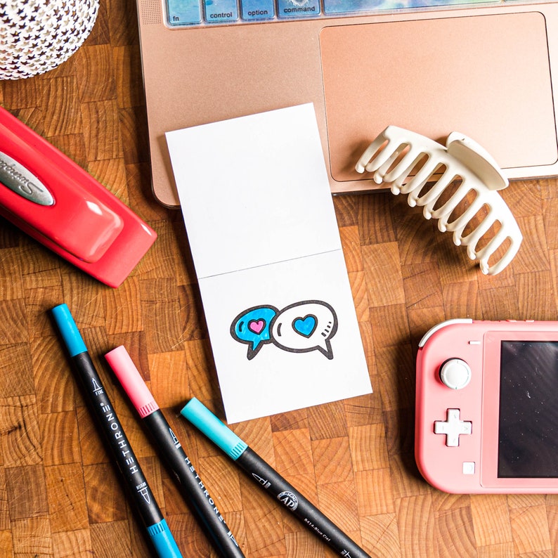 May include: A white card with a blue and pink heart design inside two speech bubbles. The card is on a wooden surface with a laptop, a stapler, and a few pens.