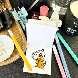 Op de afbeelding: Een klein wit notitieboekje met een cartoonillustratie van een kat op de voorkant. Het notitieboekje is omringd door verschillende items, waaronder een waterfles, een kaars, een lipgloss en een paar pennen.
