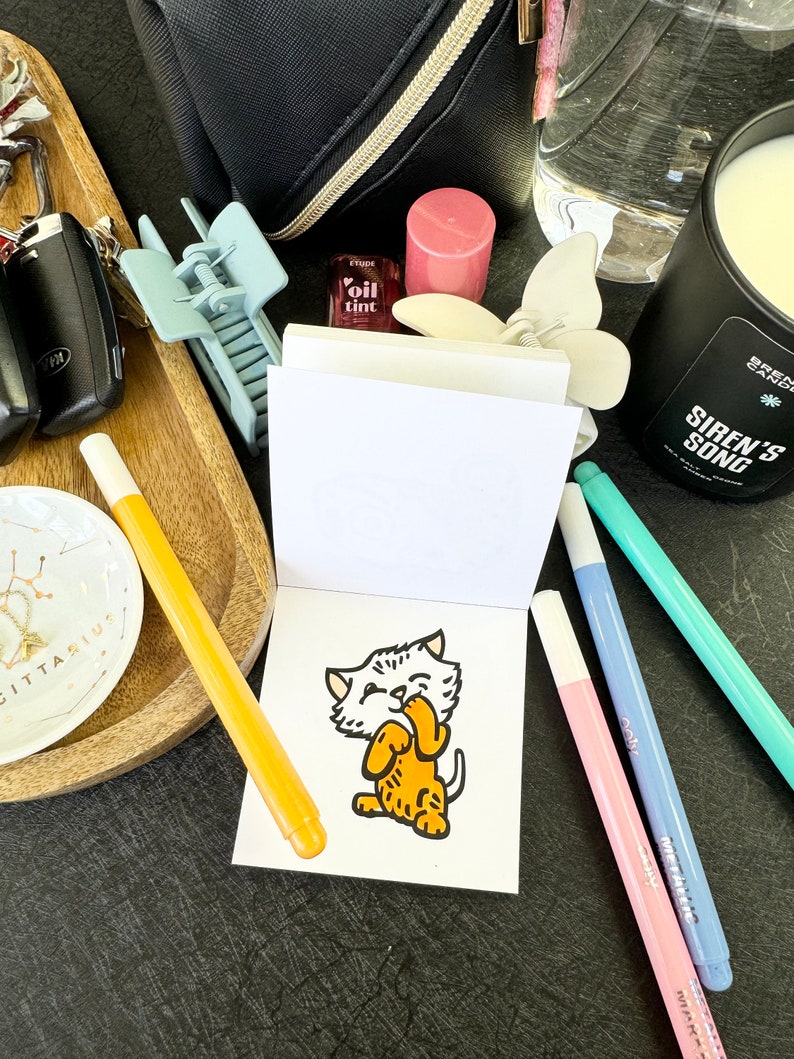 Op de afbeelding: Een klein wit notitieboekje met een cartoonillustratie van een kat op de voorkant. Het notitieboekje is omringd door verschillende items, waaronder een waterfles, een kaars, een lipgloss en een paar pennen.