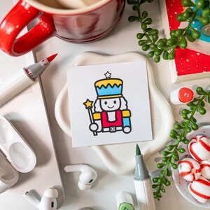 May include: A square white card featuring a colorful cartoon illustration of a nutcracker wearing a blue and yellow hat, a red jacket, and holding a yellow star-tipped wand. The nutcracker has a simple black outline and a smiling face.