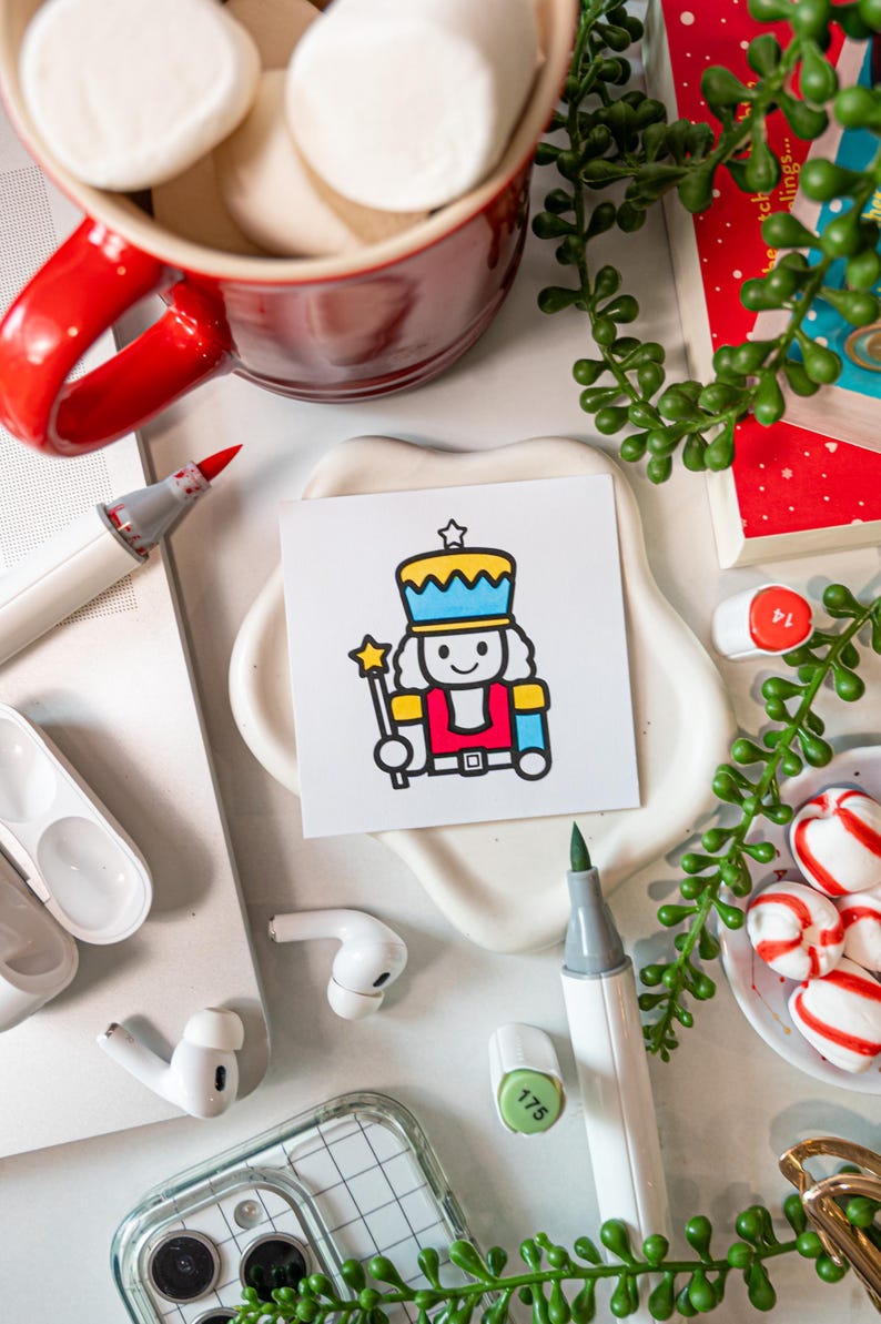 May include: A square white card featuring a colorful cartoon illustration of a nutcracker wearing a blue and yellow hat, a red jacket, and holding a yellow star-tipped wand. The nutcracker has a simple black outline and a smiling face.