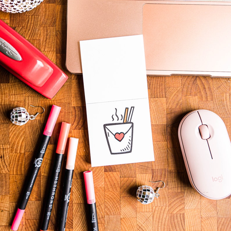 May include: A white card with a hand-drawn illustration of a steaming takeout container with chopsticks and a heart. The card is surrounded by colorful markers, a pink stapler, and a pink computer mouse with the word "logi" on it.