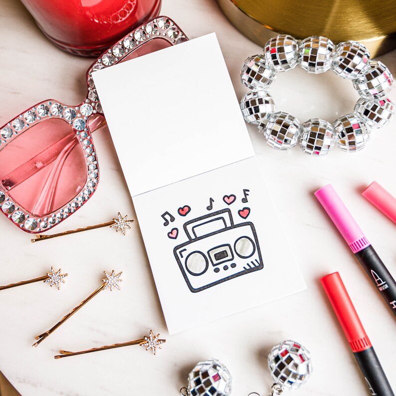 May include: A white card with a black and white drawing of a boombox with red hearts and musical notes around it. The card is surrounded by pink and red highlighters, gold hair clips, pink sunglasses with rhinestones, and silver disco balls.