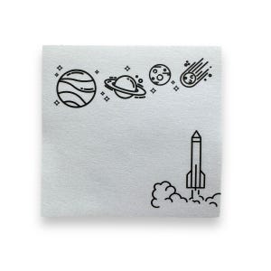 May include: A white sticky note with a black line drawing of a rocket taking off, with a cloud of smoke behind it. Above the rocket are black line drawings of planets, stars, and a comet.