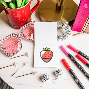 May include: A white card with a red and brown strawberry drawing on it. The card is surrounded by colorful markers, a pair of pink sunglasses, and silver disco ball earrings.