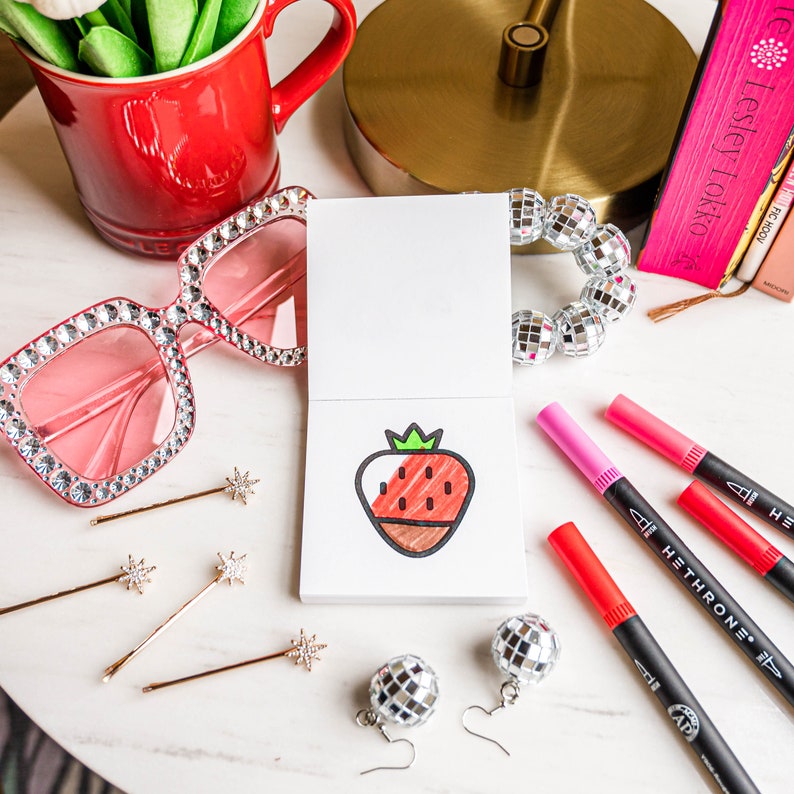 May include: A white card with a red and brown strawberry drawing on it. The card is surrounded by colorful markers, a pair of pink sunglasses, and silver disco ball earrings.