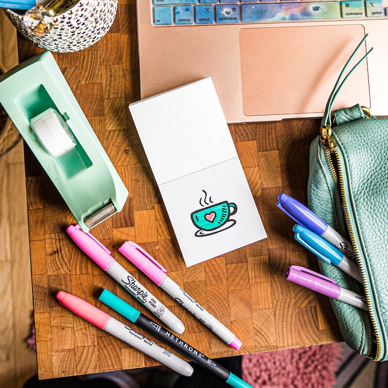 May include: A white notepad with a hand-drawn illustration of a steaming cup of coffee with a heart inside. The notepad is surrounded by a variety of colorful markers on a wooden surface.