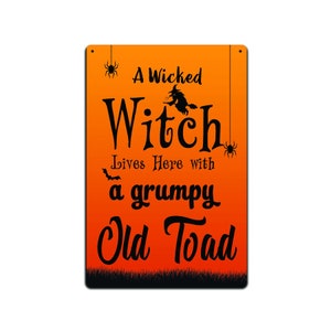 Könnte beinhalten: Ein orangefarbenes und schwarzes Halloween-Schild mit dem Text "A Wicked Witch Lives Here with a Grumpy Old Toad". Das Schild zeigt eine Hexe, die auf einem Besen fliegt, und eine Spinne.
