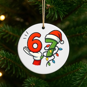 May include: A white ceramic Christmas ornament featuring the numbers 6 and 7. The number 6 is red and held by a gloved hand. The number 7 is green with a Santa hat and wrapped in colourful Christmas lights. A gold string is attached.