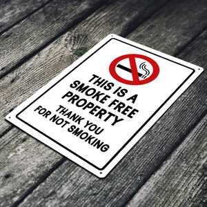 Puede incluir: Un letrero de metal blanco con texto negro que dice "This is a smoke free property. Thank you for not smoking." Un c&iacute;rculo rojo con una l&iacute;nea diagonal que lo atraviesa y un cigarrillo con humo saliendo de &eacute;l se encuentra en la esquina superior izquierda.