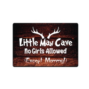 Pode incluir: Uma placa de madeira com fundo marrom rústico e texto branco. A placa diz "Little Man Cave No Girls Allowed (Except Mommy!)" com um par de chifres de veado acima do texto.