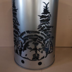 Può includere: Un braciere in metallo argentato con un designo a taglio di un muso di lupo e alberi. Il braciere è cilindrico e ha un coperchio.