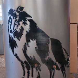 Può includere: Un focolare in metallo argentato con una silhouette nera di un cane ritagliata. Il focolare ha diversi fori intorno alla parte inferiore.