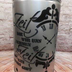 Può includere: Un focolare in metallo argentato con un design a taglio nero. Il design presenta un quadrante dell'orologio con numeri romani e le parole "Zeit die wir uns nehmen ist Zeit die uns etwas gibt".