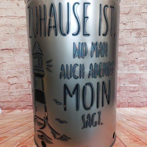 Può includere: Un bidone della spazzatura in metallo argentato con il testo tedesco "ZUHAUSE IST, WO MAN AUCH ABENDS MOIN SAGT." Un faro e gabbiani sono incisi sul lato. Il bidone ha un coperchio e dei fori.