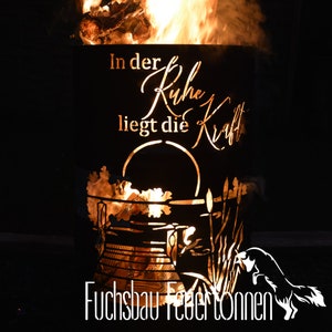 Può includere: Un braciere in metallo nero con un design a taglio di una scena lacustre e il testo "In der Ruhe liegt die Kraft" (Nella quiete risiede la forza). Il braciere è acceso e le fiamme sono visibili. Il testo "Fuchsbau Feuertonnen" è visibile nella parte inferiore dell'immagine.