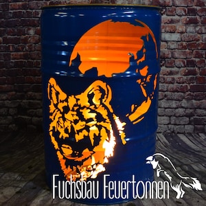 Può includere: Un barile di metallo blu con un disegno di lupo intagliato e una silhouette di luna piena, illuminato con luce arancione. Il testo "Fuchsbau Feuertonnen" è in basso.