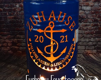 May include: A blue metal fire pit with an orange anchor design and the text "ZUHAUSE 20 21 FALLENLASSEN & LIEGENBLEIBEN". The fire pit is lit and glowing orange.