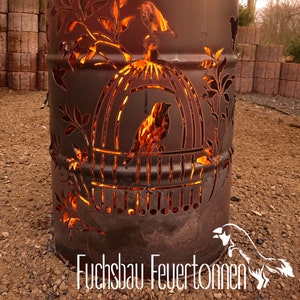 Può includere: Un focolare in metallo arrugginito con un design a taglio di uccelli e una gabbia per uccelli. Il focolare è acceso e brilla di un colore arancione. Il testo "Fuchsbau Feuertönnen" è visibile nella parte inferiore dell'immagine.