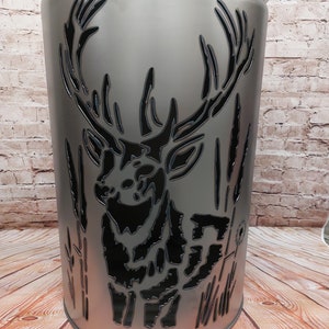 Può includere: Un braciere in metallo argentato con un design a taglio di un cervo in piedi nell'erba alta. Il braciere ha un coperchio.