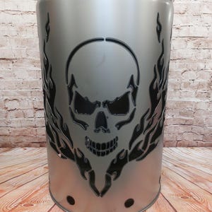 Può includere: Un bidone della spazzatura in metallo argentato con un design a teschio intagliato e fiamme. Il bidone cilindrico ha un coperchio. Il teschio è centrato, con fiamme su entrambi i lati. È posto su una superficie di legno.