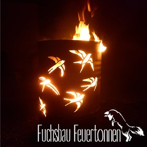 Può includere: Un braciere in metallo cilindrico con disegni di uccelli ritagliati, illuminato da fiamme. Il braciere ha una finitura scura. Le fiamme sono di colore arancione brillante e giallo, creando un bagliore caldo. Il testo "Fuchsbau Feuertonnen" è in basso.