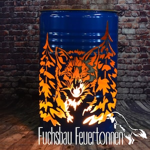 Può includere: Un barile di metallo blu con un design ritagliato di un volto di volpe circondato da alberi. Il barile è illuminato dall'interno, creando un bagliore caldo. Il testo "Fuchsbau Feuertonnen" è visibile nella parte inferiore dell'immagine.