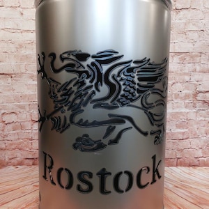 Può includere: Un braciere in metallo argentato con un designo di aquila nera e la parola "Rostock" tagliata. Il braciere ha un coperchio e dei fori sul fondo.