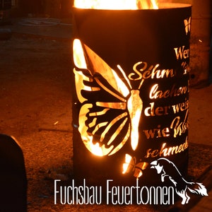 Può includere: Un braciere in metallo nero con un design a forma di farfalla tagliata. Il braciere è acceso e le fiamme sono visibili attraverso il taglio. Il testo "Wer im Leben schon lachen lernt, der weiß wie kostbar Schmerz ist" è visibile sul lato del braciere. Il testo "Fuchsbau Feuertönnen" è visibile sul fondo del braciere.