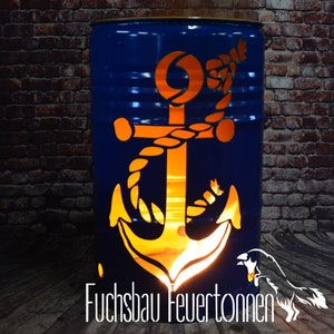 Könnte beinhalten: Ein blauer Metallfass-Feuerkorb mit einem ausgeschnittenen Anker-Design. Der Anker ist von innen beleuchtet und erzeugt ein warmes Leuchten. Der Text "Fuchsbau Feuertönnen" ist am unteren Bildrand sichtbar.