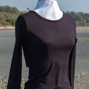 Puede incluir: Una camiseta negra de manga larga con cuello redondo. La camiseta está en un maniquí blanco y se fotografía sobre un fondo de playa.